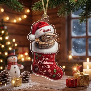 May include: A clear acrylic Christmas ornament shaped like a stocking, featuring a brown snake wearing a red and white Santa hat. The text reads Miami, First Christmas 2026, with white snowflake and green holly leaf accents on the red stocking.