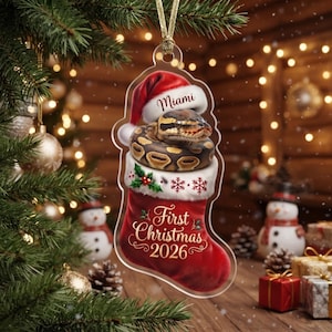 May include: A clear acrylic Christmas ornament shaped like a stocking, featuring a ball python wearing a Santa hat with the text Miami, and the text First Christmas 2026 printed in gold script on the red stocking with white fur trim.