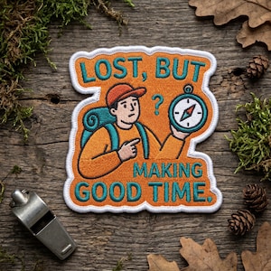 以下が含まれることがあります： 「LOST, BUT MAKING GOOD TIME」の文字が入った刺繍パッチ。バックパック、コンパス、疑問符を持った漫画のハイカーが描かれています。パッチはオレンジ、ティール、白で、白い縁取りがあります。銀色の笛が左下にあります。