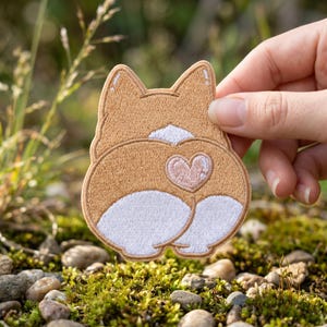 May include: An embroidered patch shaped like the rear of a Corgi dog, featuring tan and white thread colors with a small pink heart detail centered on the back. The patch has a stitched border and depicts the dog from a rear-facing perspective.