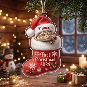 Pode incluir: Um enfeite de Natal em acrílico transparente em forma de meia, com uma cobra castanha usando um gorro de Pai Natal vermelho e o texto Miami. A meia tem rebordo de pelo branco, padrões de flocos de neve, azevinho e First Christmas 2026.