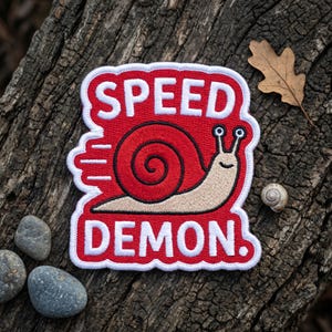 Könnte beinhalten: Gestickter Aufnäher mit rotem Hintergrund und weißem Rand. Der Aufnäher zeigt eine Cartoon-Schnecke mit den Worten "SPEED DEMON" in Weiß. Die Schnecke ist beige mit rotem Gehäuse und schwarzen Details. Der Aufnäher befindet sich auf einer verwitterten Holzoberfläche.