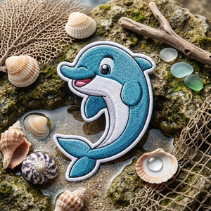 May include: An embroidered patch featuring a cartoon dolphin with a blue body and white underbelly. The dolphin is depicted in a curved, jumping pose with a smiling expression, black eyes, and a pink tongue, finished with a white stitched border.