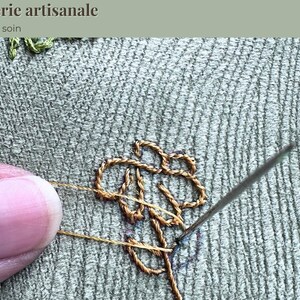 Op de afbeelding: Close-up van handborduurwerk in uitvoering. Een naald wordt in de stof gestoken, waardoor een gedetailleerd bloemmotief met gouden draad ontstaat. De tekst "Broderie artisanale, Fait avec soin" staat bovenaan.