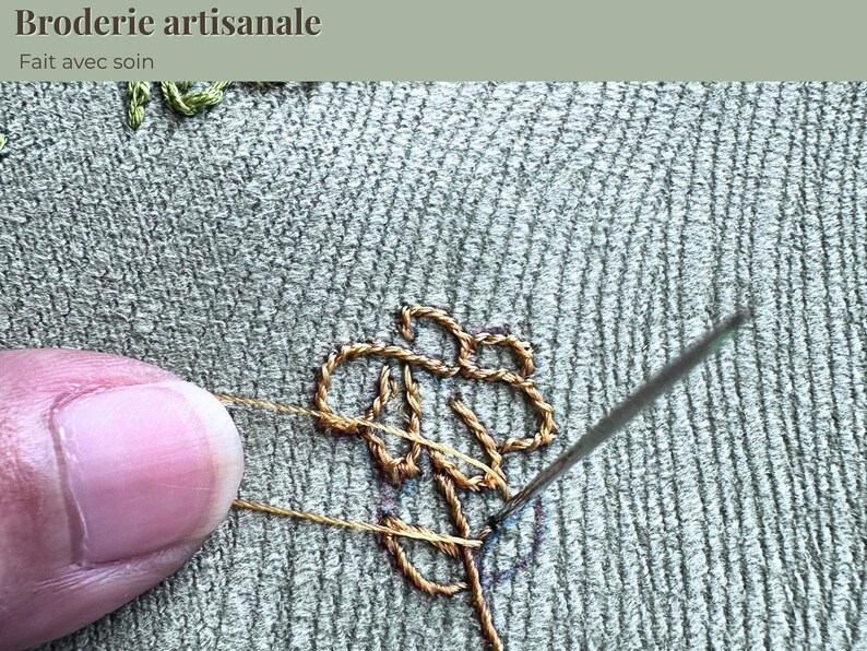 Op de afbeelding: Close-up van handborduurwerk in uitvoering. Een naald wordt in de stof gestoken, waardoor een gedetailleerd bloemmotief met gouden draad ontstaat. De tekst "Broderie artisanale, Fait avec soin" staat bovenaan.