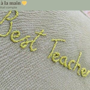 Op de afbeelding: Close-up van een lichtgroene stof met gele geborduurde tekst die luidt "Best Teacher Ever". De woorden "Brode a la main" en "Chaque detail compte" staan boven de tekst.