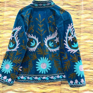 K&ouml;nnte beinhalten: T&uuml;rkisblaue Samtjacke mit aufwendiger Blumenstickerei in Braun, Hellblau und Lavendel. Die Jacke hat einen hohen Kragen und detaillierte Muster auf den &Auml;rmeln und dem R&uuml;cken, im Bohemian-Stil.