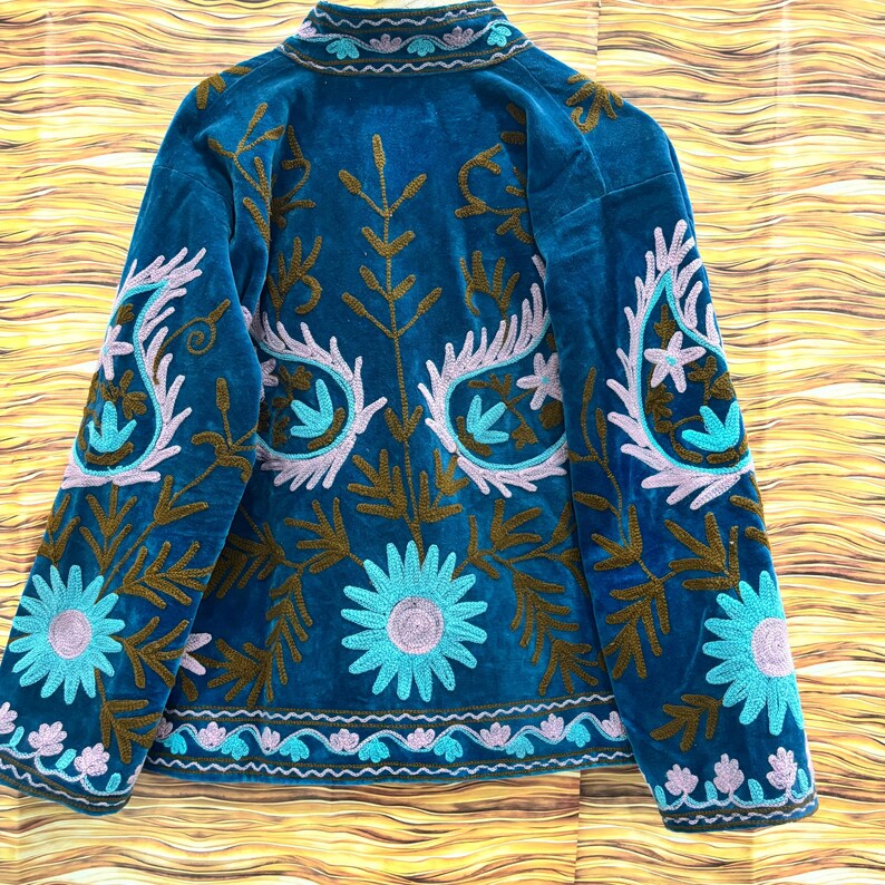 K&ouml;nnte beinhalten: T&uuml;rkisblaue Samtjacke mit aufwendiger Blumenstickerei in Braun, Hellblau und Lavendel. Die Jacke hat einen hohen Kragen und detaillierte Muster auf den &Auml;rmeln und dem R&uuml;cken, im Bohemian-Stil.