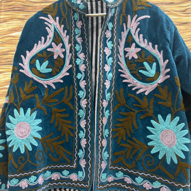 K&ouml;nnte beinhalten: T&uuml;rkisblaue Samtjacke mit aufwendiger Blumenstickerei in Rosa, Hellblau und Braun. Die Jacke hat ein Paisley-Muster und ein gestreiftes Futter, wodurch eine Boh&egrave;me-&Auml;sthetik entsteht. Die Jacke h&auml;ngt an einem Holzkleiderb&uuml;gel.
