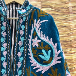 K&ouml;nnte beinhalten: T&uuml;rkisblaue Samtjacke mit aufwendiger Blumenstickerei in Hellblau, Rosa und Braun. Die Jacke hat einen hohen Kragen und ein gestreiftes Futter. Die Stickerei umfasst Paisley- und Blumenmuster.