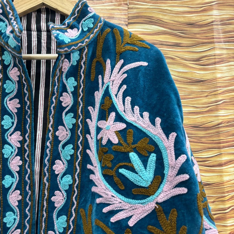 K&ouml;nnte beinhalten: T&uuml;rkisblaue Samtjacke mit aufwendiger Blumenstickerei in Hellblau, Rosa und Braun. Die Jacke hat einen hohen Kragen und ein gestreiftes Futter. Die Stickerei umfasst Paisley- und Blumenmuster.