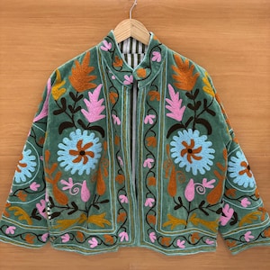 Può includere: Una giacca in velluto verde con ricami floreali intricati in arancione, rosa e blu. La giacca presenta un collo alto e una fodera a righe, creando un'estetica bohémien. Il ricamo include fiori e motivi a foglie stilizzati.