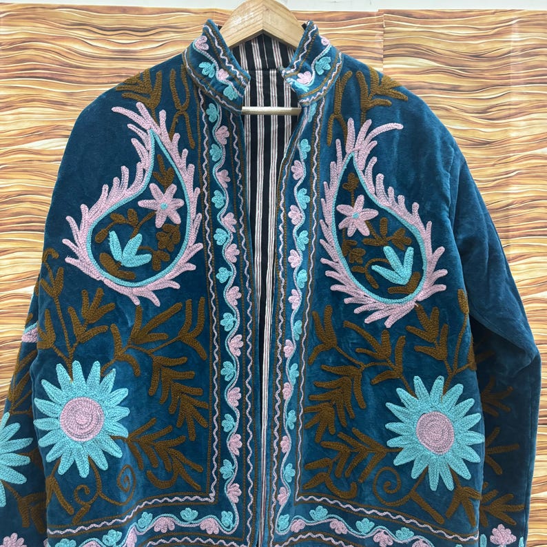 K&ouml;nnte beinhalten: T&uuml;rkisblaue Samtjacke mit aufwendiger Stickerei. Die Jacke zeigt florale und Paisley-Muster in Braun-, Rosa- und Blaut&ouml;nen. Der Kragen und die vorderen Kanten sind mit einem kontrastierenden Streifenstoff gef&uuml;ttert. Die Jacke h&auml;ngt an einem Holzkleiderb&uuml;gel.