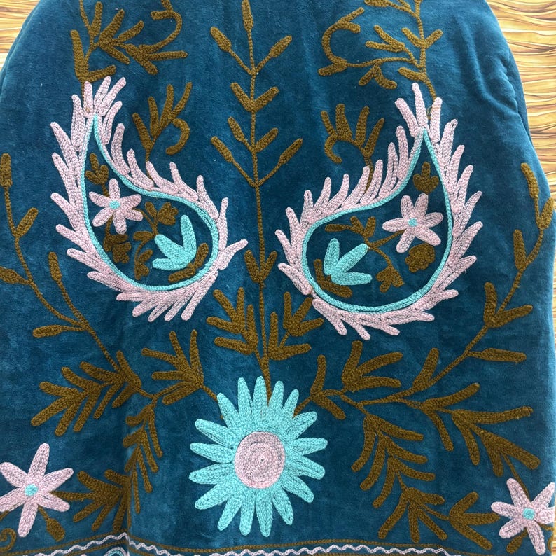 K&ouml;nnte beinhalten: T&uuml;rkisfarbener bestickter Stoff mit floralem und Paisley-Design. Die Stickerei verwendet hellrosa, t&uuml;rkis und braunes Garn. Das Design umfasst Paisley-Formen, Blumen und Blattmuster. Der Stoff hat eine weiche, samtige Textur.
