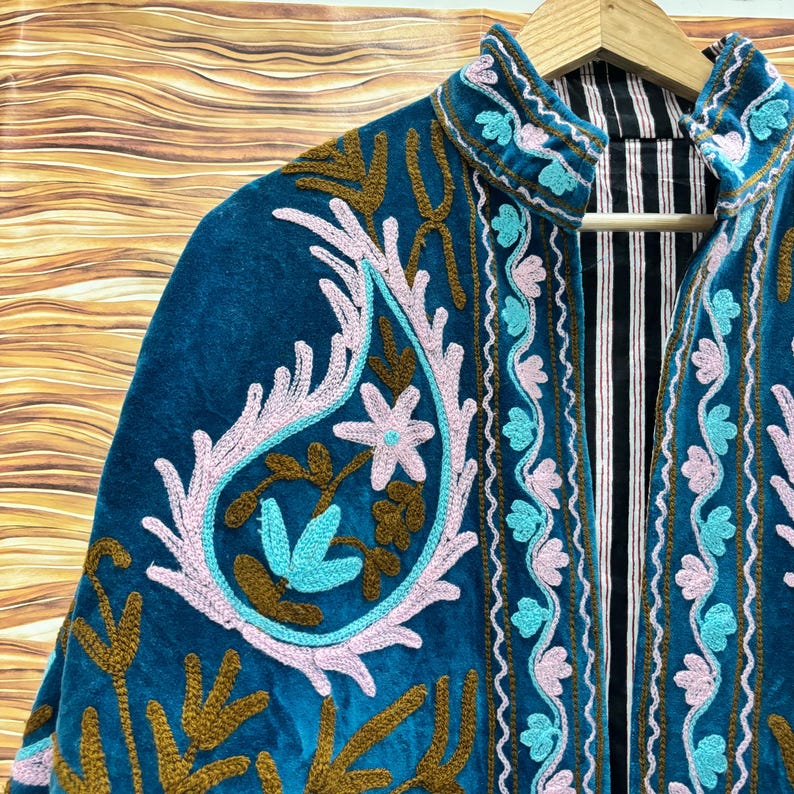 K&ouml;nnte beinhalten: T&uuml;rkisblaue Samtjacke mit aufwendiger Stickerei in Hellrosa, T&uuml;rkis und Braun. Die Jacke hat einen hohen Kragen und ein vom Paisley-Muster inspiriertes Design auf der Vorderseite. Das Innenfutter hat ein schwarz-wei&szlig;es Streifenmuster.