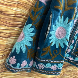 K&ouml;nnte beinhalten: T&uuml;rkisblaue Samtjacke mit floralen Stickereien. Die Jacke hat gro&szlig;e, hellblaue und rosa gestickte Blumen, braune Bl&auml;tter und dekorative Verzierungen. Das Design ist detailliert und die Jacke scheint handgefertigt zu sein.