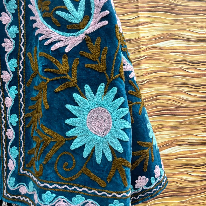 K&ouml;nnte beinhalten: Nahaufnahme eines tealfarbenen Samttextils mit aufwendiger Stickerei. Das Design zeigt gro&szlig;e t&uuml;rkisfarbene und rosa Blumenmuster sowie braune blattartige Formen. Die R&auml;nder sind mit einem wei&szlig;en und t&uuml;rkisfarbenen Wellenrand versehen.