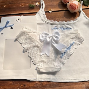 Può includere: Un completo intimo bianco, composto da una canotta e biancheria intima con bordi in pizzo, legato con un fiocco bianco. La canotta e la biancheria intima presentano un testo ricamato blu. Il set è presentato con una confezione regalo e rose.