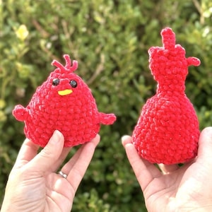 May include: Two handmade red crochet figures. One is a round creature with a yellow beak and black eyes. The other is a figure of a hand with the middle finger extended. The background is green foliage.