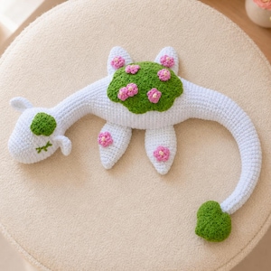 Könnte beinhalten: Ein weißes gehäkeltes Dinosaurier-Spielzeug mit langem Hals und Schwanz. Die Kreatur hat einen grünen Fleck auf Rücken und Kopf, der mit kleinen rosa gehäkelten Blumen verziert ist. Weitere rosa Blumen befinden sich an den Flossen und am Schwanz.
