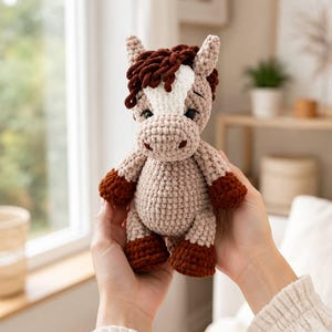May include: A crochet horse stuffed animal featuring a light tan body, dark brown hooves, and a dark brown mane. The horse has a white patch on its forehead, small pointed ears, and black safety eyes, crafted with a soft, textured yarn finish.