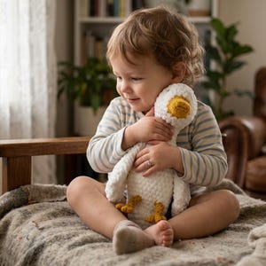 Könnte beinhalten: Ein Kind hält ein gehäkeltes weißes Gänse-Plüschtier mit gelbem Schnabel und Füßen. Die Gans hat einen strukturierten Körper. Das Kind trägt ein gestreiftes Oberteil und sitzt auf einer Decke.