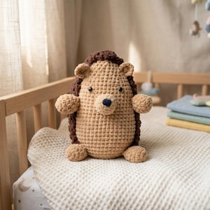 Puede incluir: Un juguete de peluche de erizo de ganchillo hecho a mano en beige y marrón, con una nariz azul oscuro y pequeños ojos negros. El animal de peluche está sentado en una manta blanca de tejido de gofre dentro de una cuna de madera. El erizo tiene brazos y piernas pequeños.