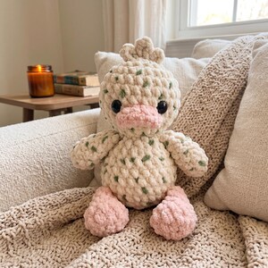 May include: A handmade crochet duck plush toy. The duck is cream-colored with green speckles, black eyes, and a pink beak and feet. It is sitting on a beige knitted blanket and pillow.