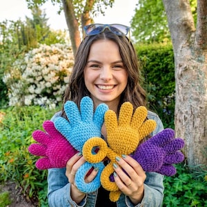 May include: A woman holds a collection of colorful, crocheted hand-shaped toys. The toys are in shades of pink, blue, yellow, and purple. The background features lush greenery and a tree.