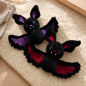 May include: Two handmade crochet bats with black yarn bodies and wings. One bat features purple inner wing and ear accents with green eyes, while the other bat has red inner wing and ear accents with pink eyes. Both bats have white fangs.