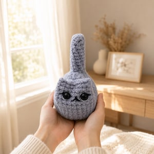 May include: A small, light grey crocheted plush toy shaped like a cat with a single upright tail. The figure features a rounded body, two small pointed ears, and two large, glossy black plastic eyes with stitched details for the nose and mouth.