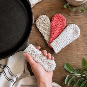 May include: A black cast iron skillet handle covered by a textured, off-white crocheted sleeve. Three additional crocheted handle covers in beige, coral, and off-white are arranged in a fan shape nearby, featuring a thick, durable yarn construction.