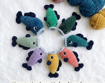 May include: A collection of handmade crochet fish in various colors, including blue, green, purple, and yellow, arranged in a circle. Each fish has dark blue fins and a white mouth. The text "Create in Any Color You Love" is at the bottom.