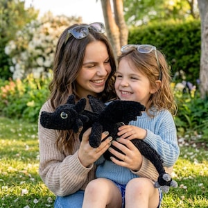 May include: A black crocheted dragon toy with green eyes and a long tail. The dragon is held by two people. The dragon has wings and a detailed head. The background is a green lawn and trees.