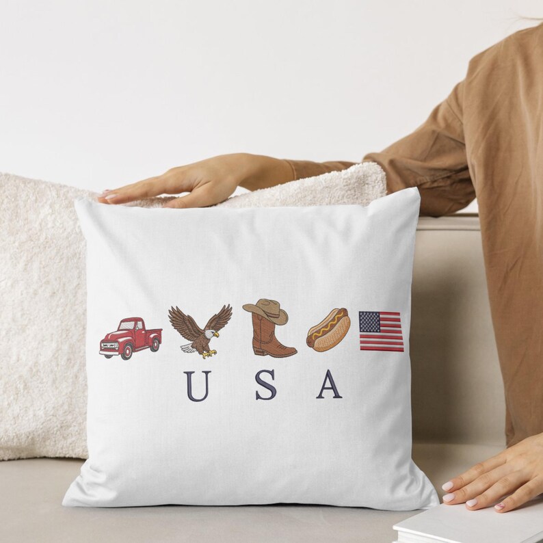 K&ouml;nnte beinhalten: Ein wei&szlig;es quadratisches Dekokissen mit einem Grafikdruck, der einen roten Pickup, einen Wei&szlig;kopfseeadler, einen braunen Cowboystiefel, einen Hotdog und die amerikanische Flagge in einer Reihe &uuml;ber den dunkelblauen Buchstaben U S A zeigt.
