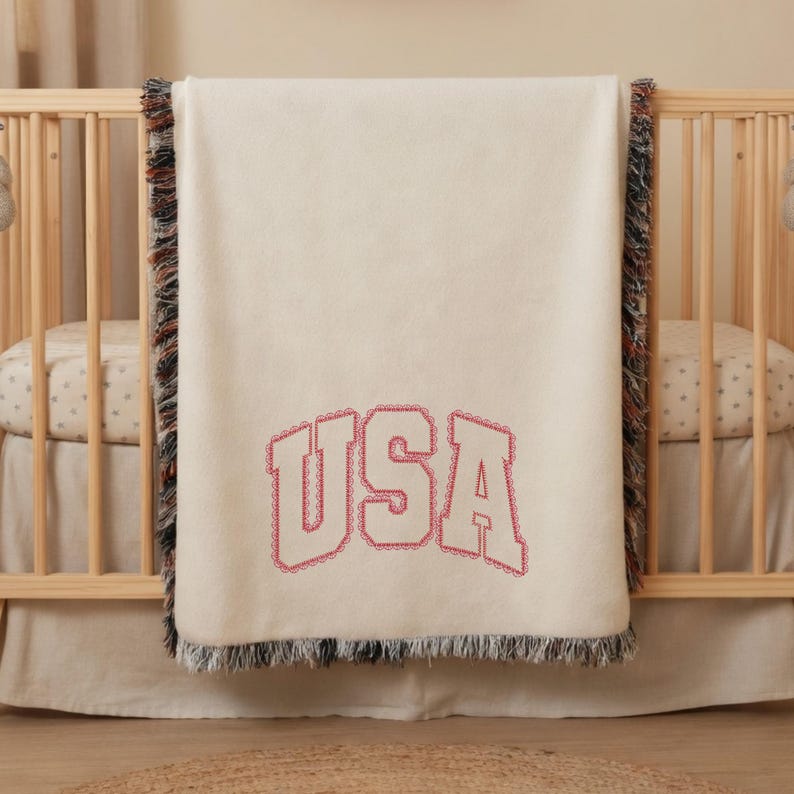 K&ouml;nnte beinhalten: Eine cremefarbene, gewebte Wohndecke mit dem Schriftzug USA in einer gro&szlig;en, roten Schriftart im College-Stil. Die Decke hat dekorative Fransenr&auml;nder mit einer Mischung aus roten, wei&szlig;en und blauen F&auml;den an den vertikalen Seiten des Stoffes.