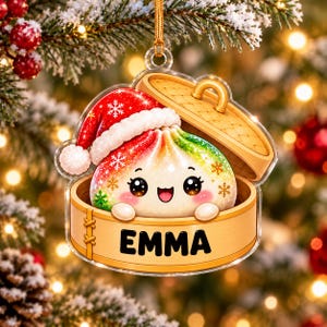 Könnte beinhalten: Ein transparenter Acryl-Weihnachtsanhänger mit einem bunten, rot-grün verlaufenden Cartoon-Dumpling, der eine rote Weihnachtsmütze mit weißem Rand trägt. Der Dumpling sitzt in einem hölzernen Dämpfkorb mit dem in Schwarz gedruckten Namen EMMA.