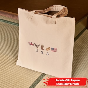 K&ouml;nnte beinhalten: Eine beigefarbene Tragetasche aus Segeltuch mit gestickten Symbolen eines roten Lastwagens, eines Adlers, eines Cowboystiefels, eines Hotdogs und der amerikanischen Flagge sowie den dunkelblau gestickten Buchstaben U S A.