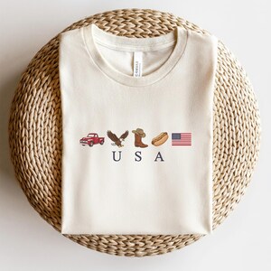K&ouml;nnte beinhalten: Ein cremefarbenes Kurzarm-T-Shirt mit einem Grafikdruck, der einen roten Pickup, einen Wei&szlig;kopfseeadler, einen braunen Cowboystiefel, einen Hotdog und die amerikanische Flagge zeigt, mit den dunkelblauen Buchstaben U S A darunter.