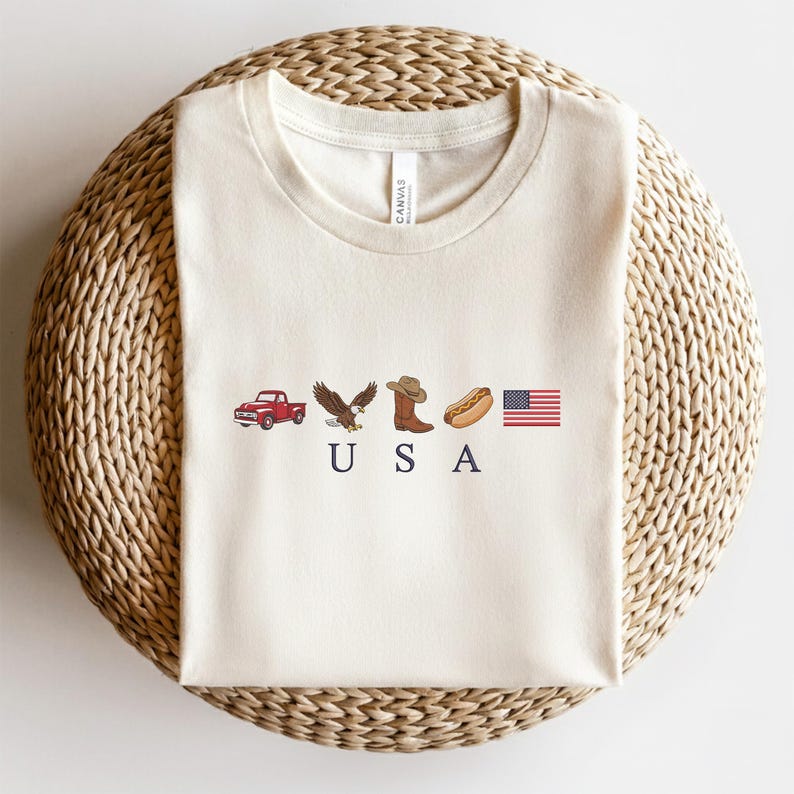 K&ouml;nnte beinhalten: Ein cremefarbenes Kurzarm-T-Shirt mit einem Grafikdruck, der einen roten Pickup, einen Wei&szlig;kopfseeadler, einen braunen Cowboystiefel, einen Hotdog und die amerikanische Flagge zeigt, mit den dunkelblauen Buchstaben U S A darunter.