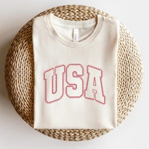 K&ouml;nnte beinhalten: Ein cremefarbenes Kurzarm-T-Shirt mit dem Text USA in einer fetten, geschwungenen College-Schriftart. Die Buchstaben sind mit einem dekorativen roten Spitzenmuster umrandet. Das Shirt hat einen Rundhalsausschnitt und eine Standardpassform.