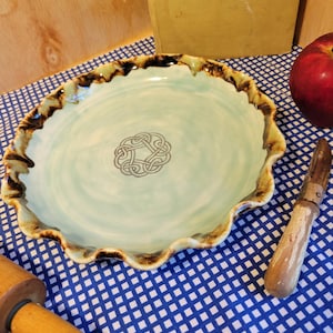 Könnte beinhalten: Eine handgefertigte Keramik-Kuchenplatte mit hellgrünem Innenraum und einem braunen und beigefarbenen, gerüschten Rand. Die Platte hat ein keltisches Knotendesign in der Mitte. Ein Kochbuch, Apfel, Nudelholz und Messer sind im Hintergrund.