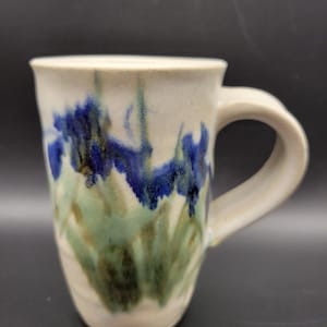 Könnte beinhalten: Ein Keramikbecher mit cremefarbener Basis und einem handgemalten Aquarell-Design aus blauen Irisblüten und grünem Blattwerk. Der Becher verfügt über einen dicken, geschwungenen Henkel und einen glatten, zylindrischen Körper mit leichtem Rand.