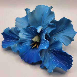 May include: A decorative flower ornament featuring layered, ruffled petals in varying shades of vibrant blue. The center of the bloom displays a pale green stamen surrounded by thin, yellow-tipped filaments, creating a detailed floral sculpture.