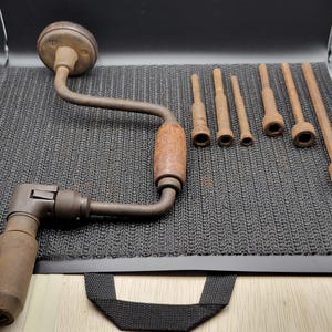 以下が含まれることがあります： 木製のハンドルと回転ノブを備えたヴィンテージの金属製ハンドドリルと、長さや直径が異なる6本の金属製ドリルビットのセット。全体的に風化した酸化した茶色の仕上げが施されています。