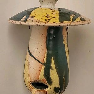 May include: A ceramic mushroom-shaped birdhouse with a speckled white top and a wide, flat, multi-colored cap. The body is a mix of yellow, green, and peach tones with a small opening and a beak-like projection. A gold chain hangs from the top.