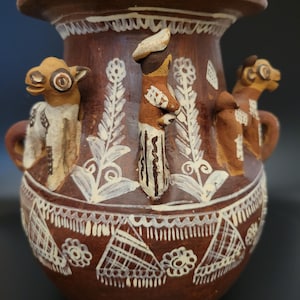 May include: A brown ceramic vase with a wide rim and two small handles. The vase is decorated with white floral and geometric patterns. Three figures are sculpted on the vase, and the vase has a dark background.