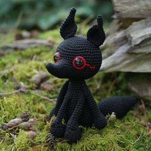 May include: A handmade black crochet fox doll with red accents around the eyes. The fox has large, pointed ears and a long tail. It is sitting on a bed of green moss and leaves.