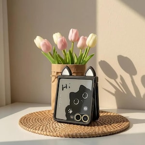 Puede incluir: Funda de teléfono negra con detalles de orejas de gato, con un diseño de gato negro y la palabra "Hi:". La funda está sobre una estera tejida, con un jarrón de tulipanes rosas y blancos en el fondo.