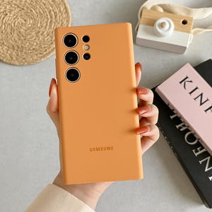 Può includere: Custodia per telefono arancione con tre obiettivi della fotocamera e il logo Samsung. La custodia è tenuta in mano con lunghe unghie arancioni. Una piccola fotocamera in legno e un libro sono sullo sfondo.